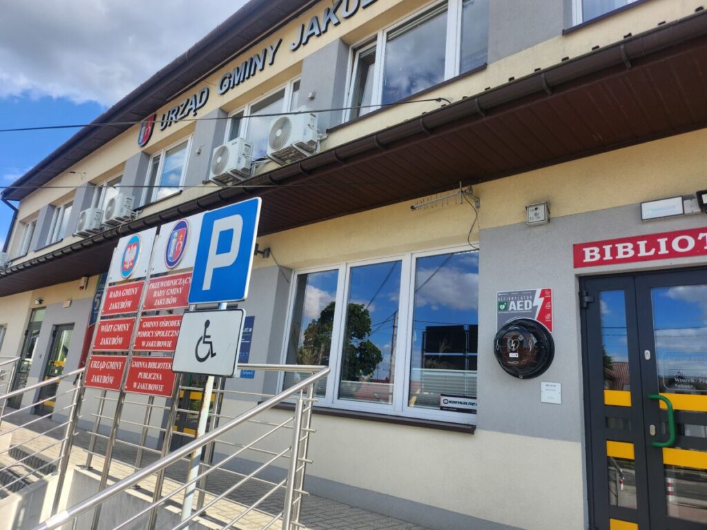 Budynek urzędu gminy z jasną fasadą. Na pierwszym planie znaki parkingowe i tablice informacyjne. Obok drzwi znajduje się defibrylator.