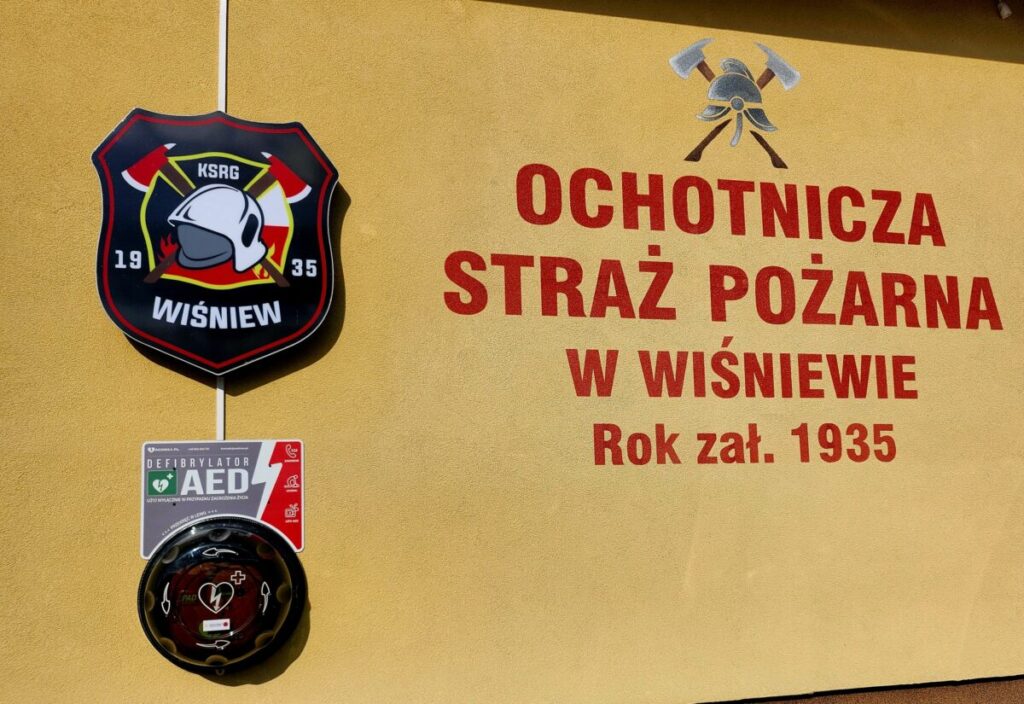Na żółtej ścianie widoczny jest herb straży pożarnej i napis Ochotnicza Straż Pożarna w Wiśniewie, rok zał. 1935. Obok znajduje się defibrylator AED. Kolory to czerwony, czarny i biały.