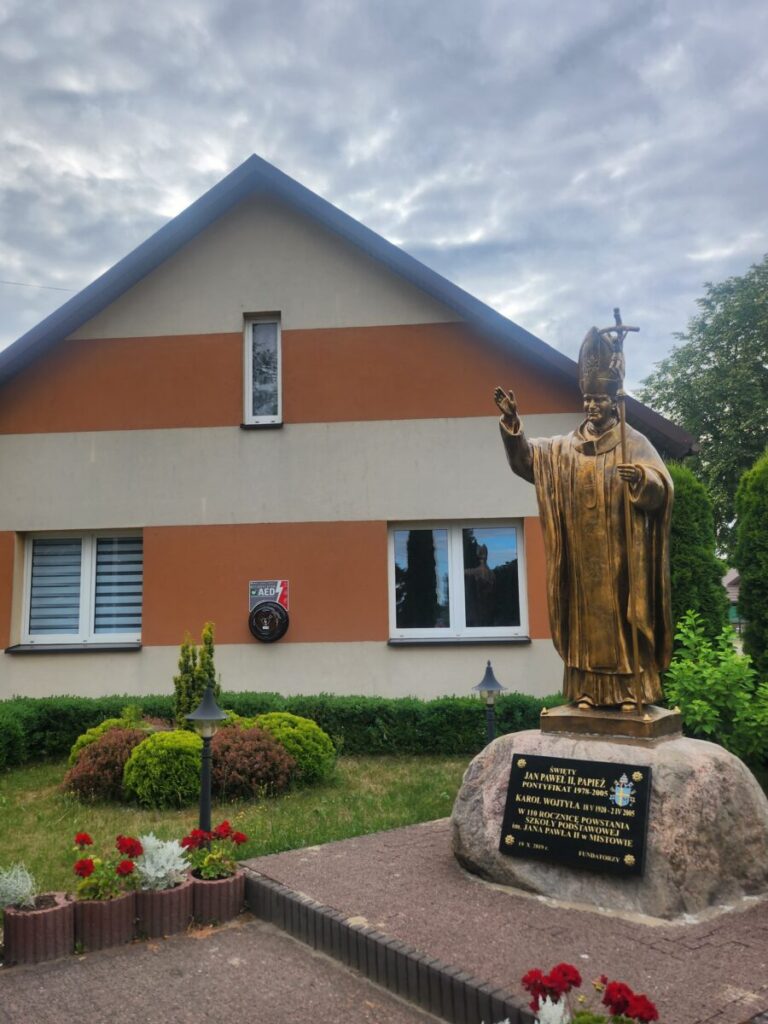 Na zdjęciu znajduje się złoty pomnik postaci na tle domu w odcieniach pomarańczowo-kremowych. Przed domem widoczne są rośliny oraz kwiaty. Niebo jest pochmurne, tworząc stonowane tło.