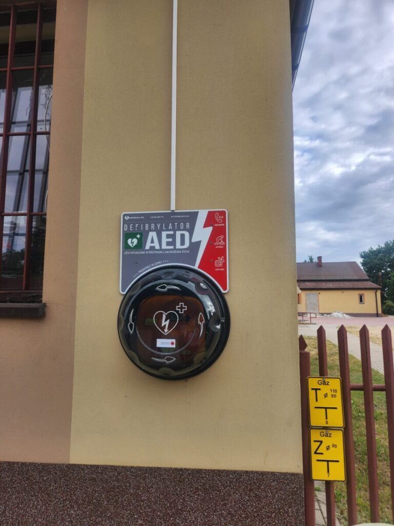 Ściana budynku z zewnątrz w odcieniach beżu. Na niej zamontowany jest defibrylator AED w czarnym, okrągłym pojemniku. Obok znajduje się biało-czerwona tablica informacyjna.
