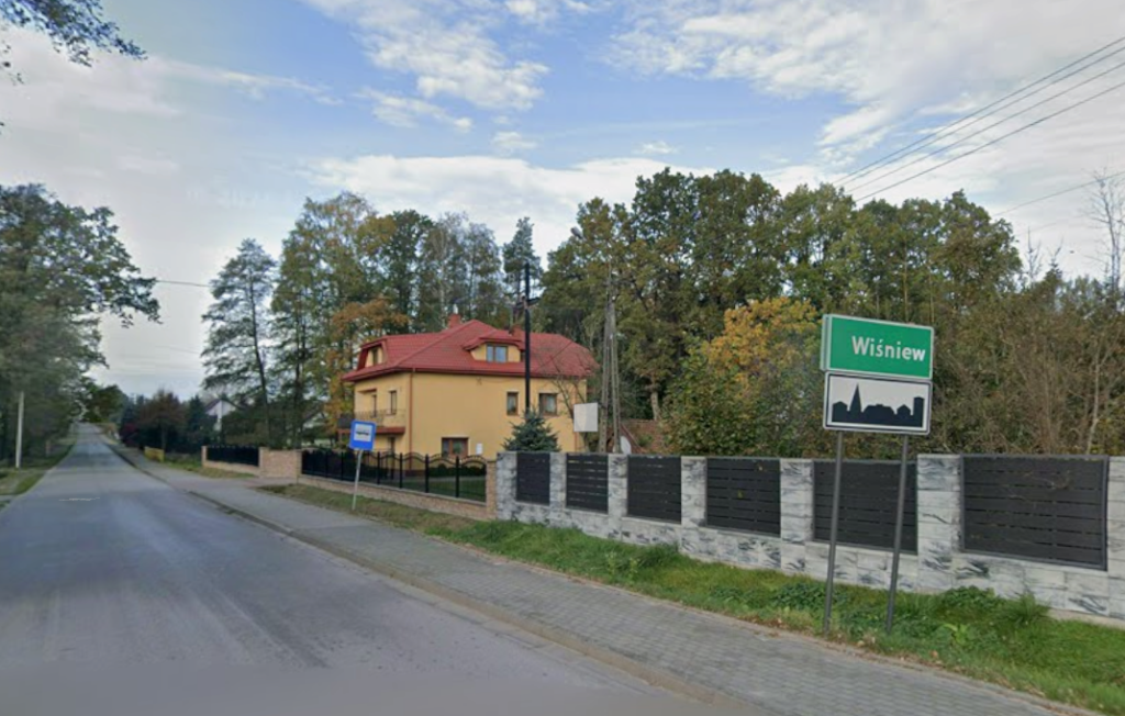 Droga z drzewami po bokach, dom w tle z czerwoną dachówką. Tablica z nazwą miejscowości Wiśniew na zielonym tle. Jasne niebo z chmurami.