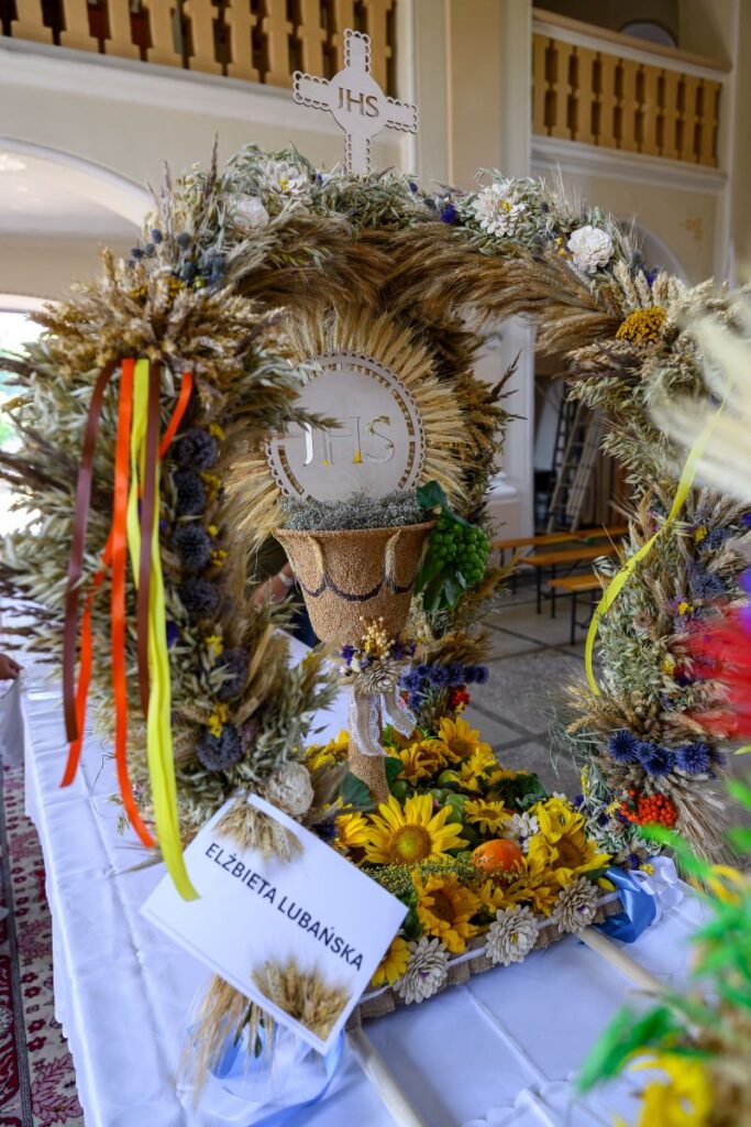 Wieniec zbożowy z kwiatami, krzyżem i napisem JHS na białym tle. Kolory: żółty, zielony, niebieski. Stół przykryty białym obrusem.