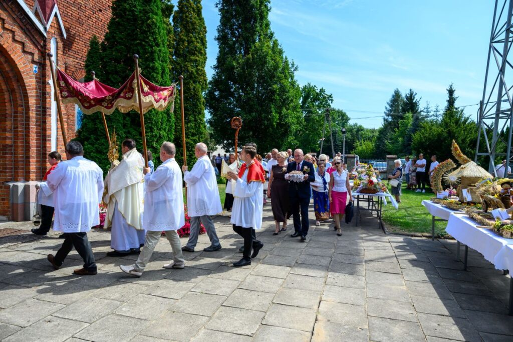 Procesja kościelna w białych szatach z czerwonym baldachimem. Tło to ceglana ściana oraz zielone drzewa. Po prawej wystawione dekoracje dożynkowe.