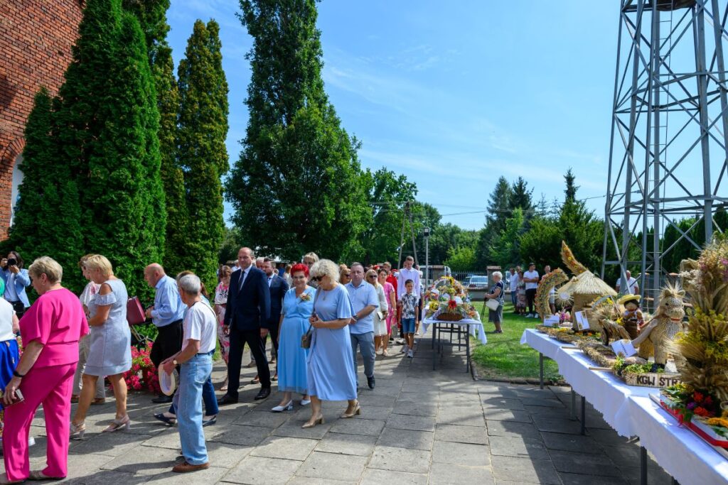 Na zdjęciu widać grupę ludzi idących po brukowanej ścieżce w otoczeniu zielonych drzew. Po prawej stronie ustawione są stoły z dekoracjami wykonanymi z ziaren i słomy. W tle znajduje się wieża stalowa na tle jasnego nieba.