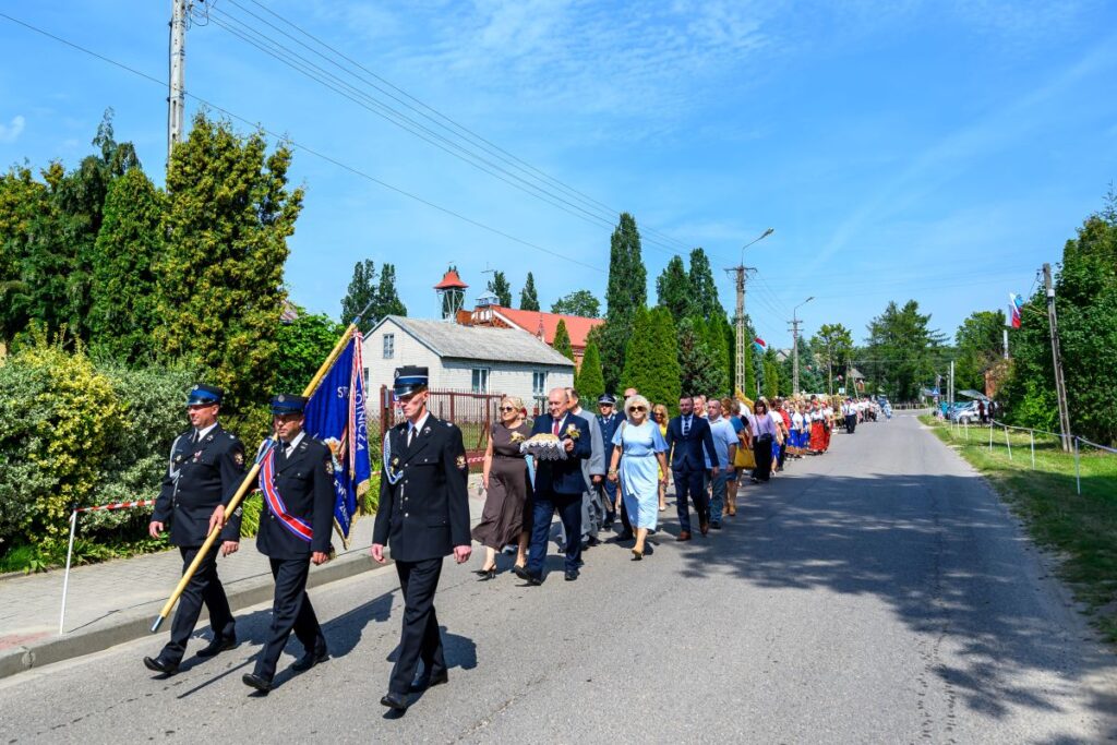Strażacy w czarnych mundurach maszerują z flagą po wiejskiej drodze. W tle drzewa i budynki o czerwonych dachach. Pogoda jest słoneczna.