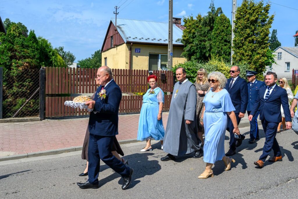 Grupa ludzi w formalnych strojach maszeruje ulicą. Mężczyzna na przedzie niesie dekoracyjne pieczywo. Tło to dom i drzewa przy ulicy.