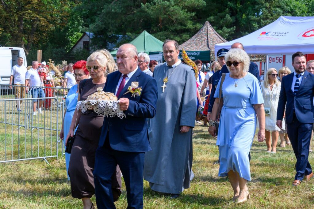 Grupa ludzi w eleganckich strojach na zewnątrz. Starszy mężczyzna niesie ozdobny chleb w otoczeniu zieleni. Tło z namiotami i trawą.