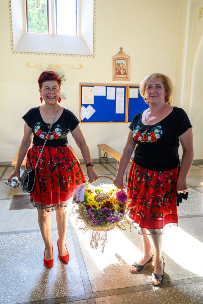 Dwie kobiety w czerwonych, kwiecistych spódnicach stoją w pomieszczeniu. Jedna z nich trzyma kosz z kolorowymi kwiatami. W tle jasne ściany i tablica.