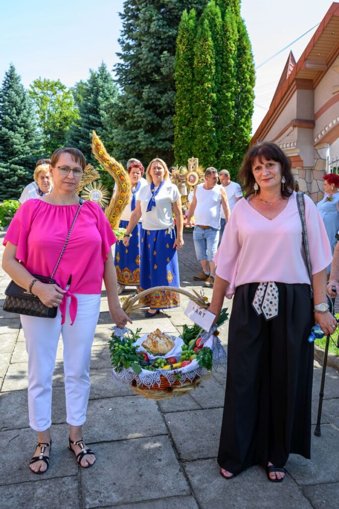 Grupa ludzi z koszem dożynkowym pozuje na tle zielonych drzew i budynku. Dwie kobiety na pierwszym planie noszą różowe bluzki. Kosz ozdobiony jest zbożem i owocami.