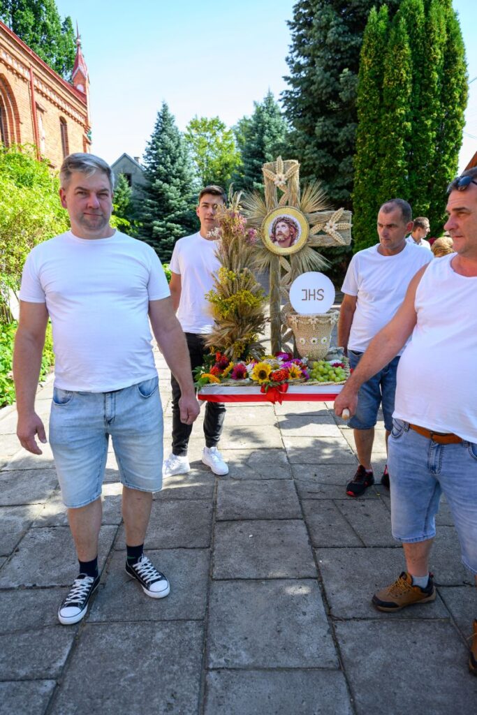 Czterech mężczyzn w białych koszulkach niesie ozdobny wieniec ze zboża i kwiatów z symbolem religijnym. Tło stanowi ceglany budynek i zielone drzewa. Na wieńcu widoczne są słoneczniki i napis J