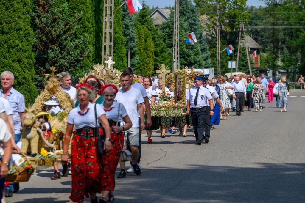 Ludzie w strojach regionalnych maszerują podczas procesji. W tle zielone drzewa i domy. Niosą ozdoby ze słomy i kwiatów.
