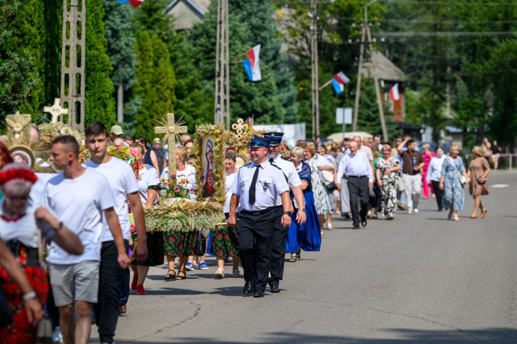 Grupa ludzi w tradycyjnych strojach idzie ulicą; niosą ozdobne wieńce. W tle drzewa i budynki. Słoneczny dzień.