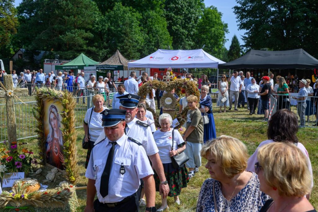 Na zdjęciu widać tłum ludzi podczas plenerowego wydarzenia z kolorowymi namiotami w tle. W centralnej części przechodzą strażacy i osoby niosące dekoracje z zboża. Obok stoi obraz Matki Boskiej ozdobiony kwiatami i zbożem.