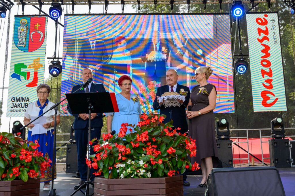 Na scenie stoi pięć osób obok dużego ekranu. Ubrani są w eleganckie stroje, a na pierwszym planie czerwone kwiaty. Po bokach widoczne są banery z napisami i herbem gminy.