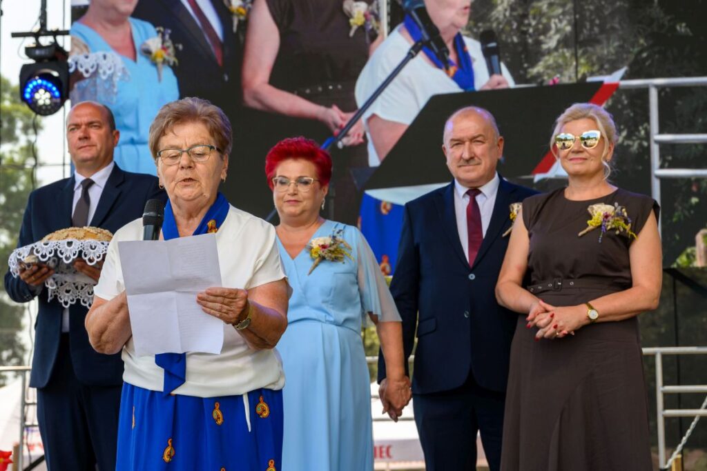 Grupa ludzi stoi na scenie; kobieta czyta z kartki. Kolory na ubraniach to niebieski, brązowy i czarny. Tło: zielone drzewa.