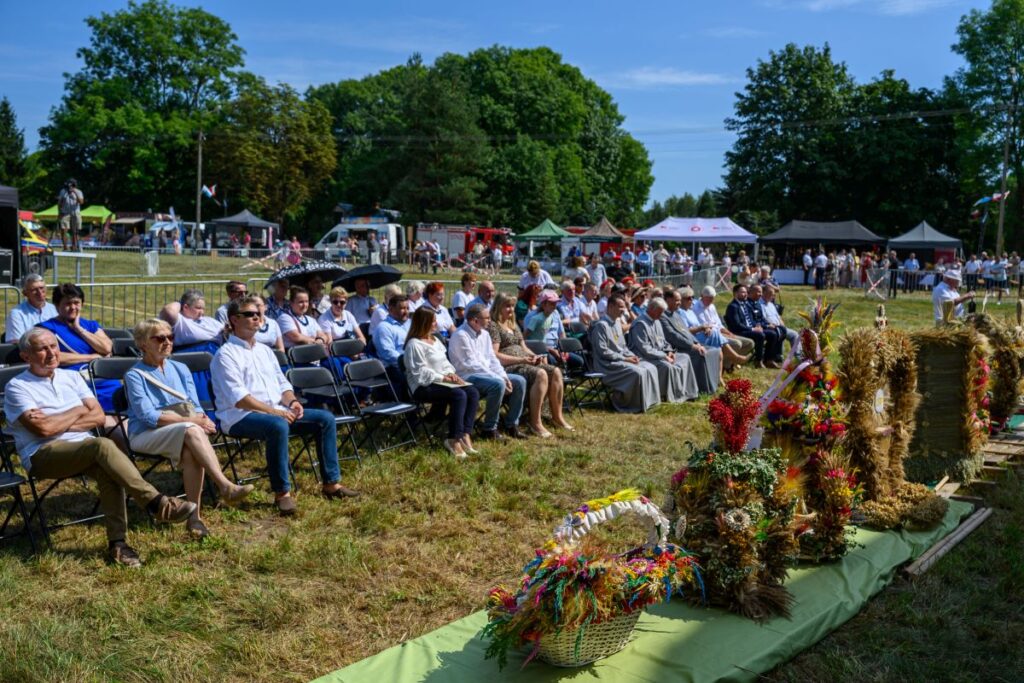 Na zdjęciu jest grupa ludzi siedząca na zewnątrz przy pięknej pogodzie. Przed nimi są ozdobne kompozycje z kwiatów i słomy. W tle widoczny jest zielony park i namioty.