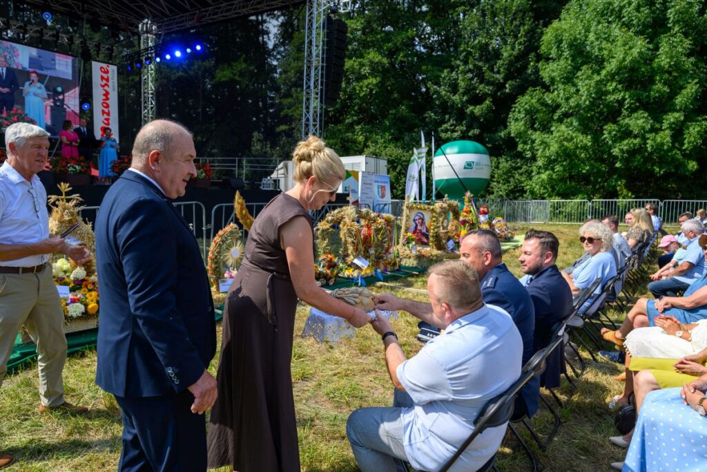 Na zdjęciu widoczna jest kobieta w ciemnej sukience wręczająca chleb siedzącym mężczyznom na zielonej trawie. Za nimi znajduje się scena z dużym ekranem i napisem Mazowsze. W tle są krzewy, dekoracje dożynkowe i bal