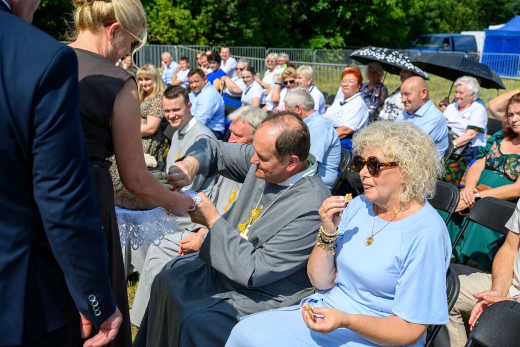 Na zdjęciu widać grupę ludzi siedzących na świeżym powietrzu podczas uroczystości. Osoba w szarym stroju podaje chleb kobiecie w niebieskiej sukience. W tle tłum, zielone drzewa i namioty.