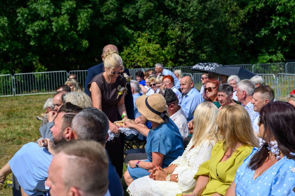 Grupa ludzi siedzi na zewnątrz na krzesłach. Kobieta w ciemnej sukience podaje jedzenie. W tle zielone drzewa i metalowe ogrodzenie.