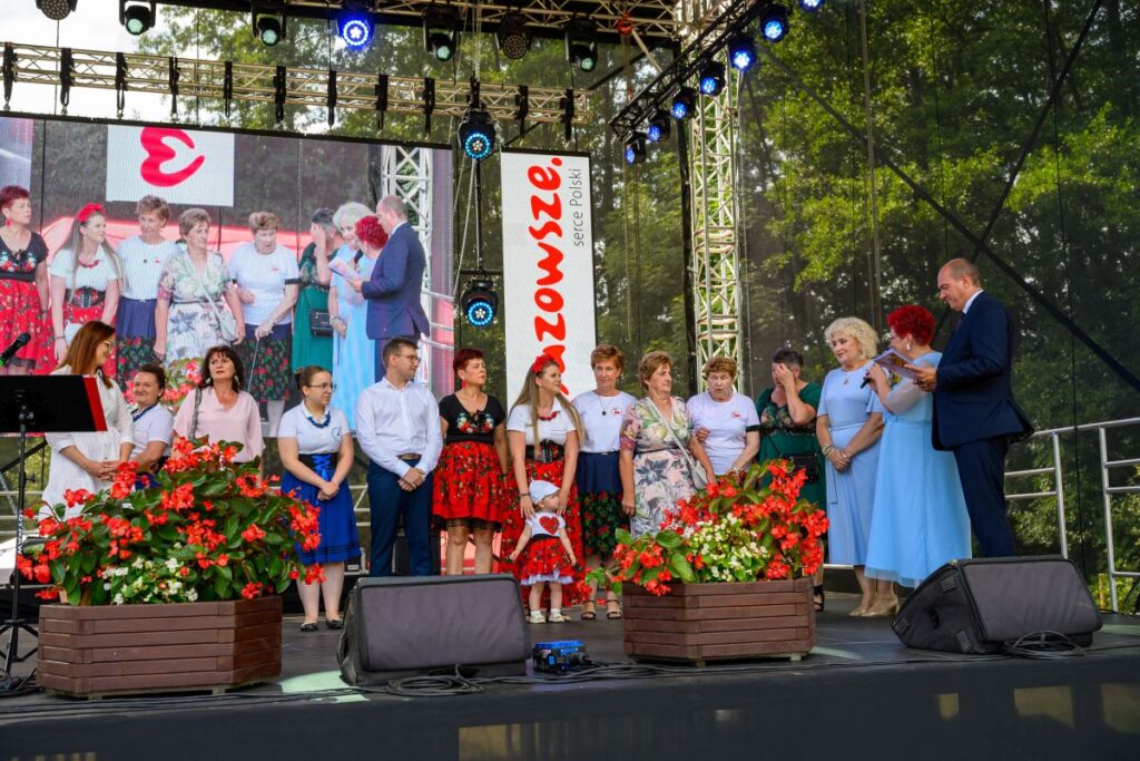 Na scenie grupa ludzi stoi przed dużym ekranem. Dominują kolory czerwony i biały, a tło to zielone drzewa. Na przodzie są kwiaty w donicach.