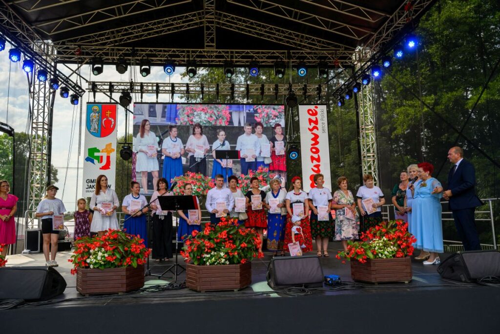 Grupa osób stoi na scenie pod zadaszeniem, trzymając dyplomy. Przed nimi czerwone kwiaty w donicach, a w tle duży ekran. Na scenie widoczne są różnokolorowe światła i napisy Mazowsze.