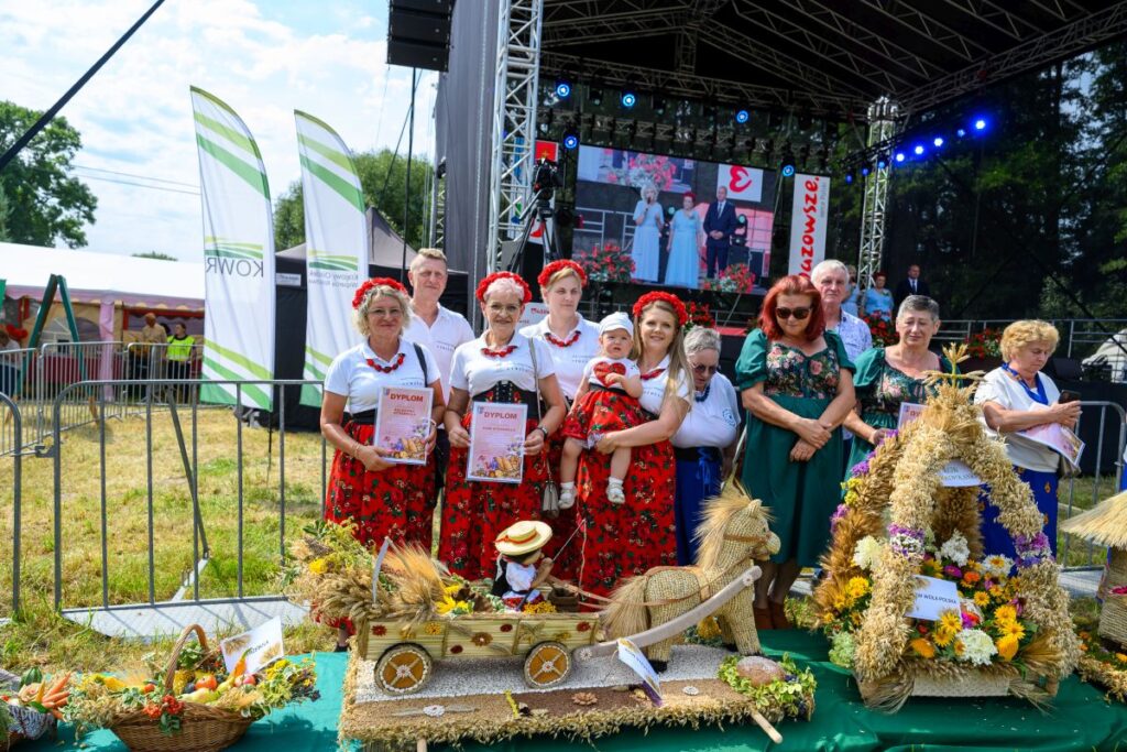 Grupa ludzi w strojach ludowych stoi przed sceną plenerową. Wokół kwiatowe kosze i ozdoby ze zboża. W tle widać ekran z kolorowymi światłami.