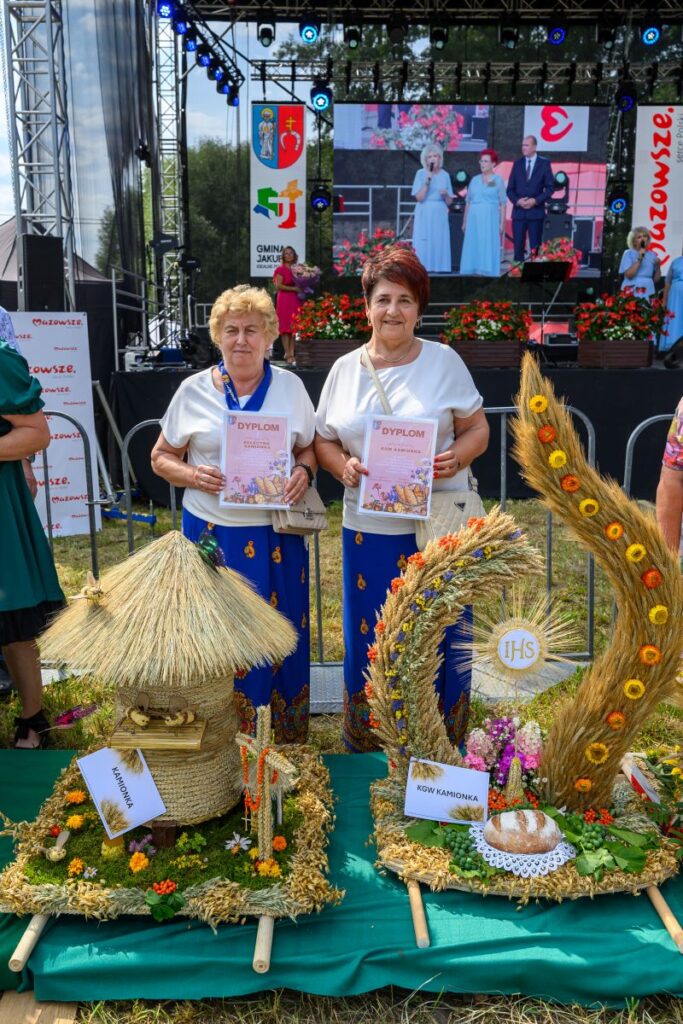 Dwie kobiety trzymają dyplomy, za nimi scena z ekranem. Przed nimi rękodzieło zbożowe. Wszystko w letnich kolorach.