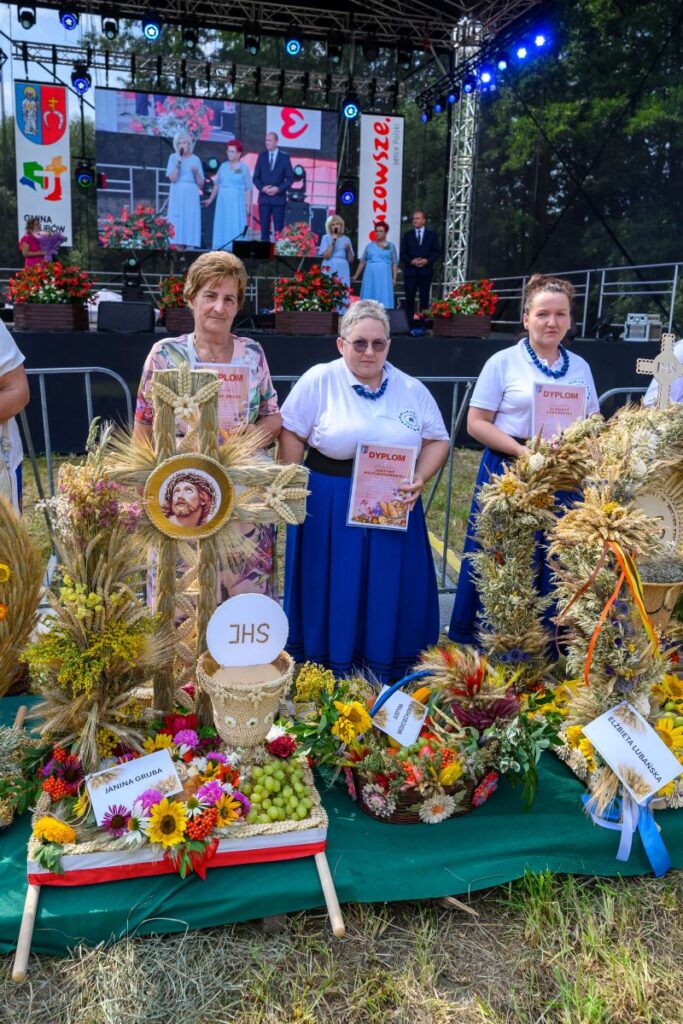 Trzy kobiety trzymają dyplomy, stoją przy dekoracjach z zbóż i kwiatów. Scena z ekranem w tle. Kolory żywe, zielone tło.