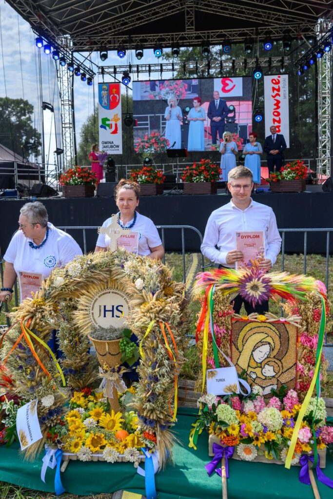Obraz przedstawia scenę z wydarzenia plenerowego. Na pierwszym planie osoby trzymają dyplomy i wieńce z kwiatów w kolorach żółtym i zielonym. W tle scena z dużym ekranem i czerwonymi kwiatami.