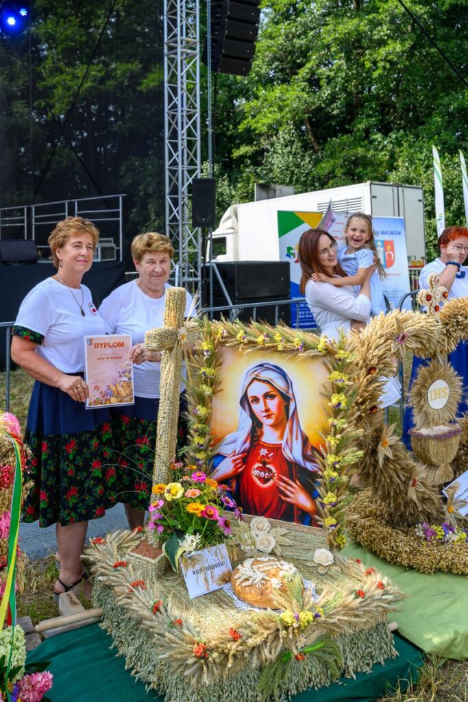 Kobiety prezentują ozdobny wieniec z wizerunkiem Matki Boskiej, otoczony kwiatami i kłosami zbóż. Tło to zielone drzewa i scena. Dziecko i kobieta po prawej dodają atmosfery radości.