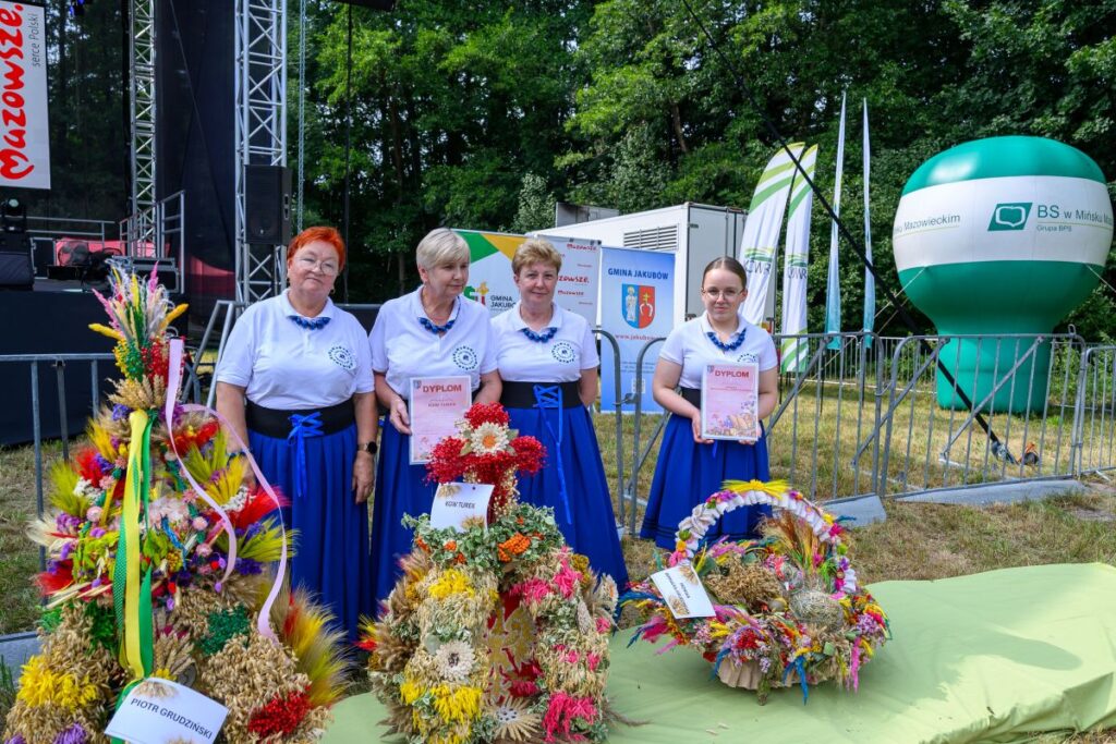 Cztery kobiety w białych bluzkach, trzymające dyplomy, stoją obok kolorowych wieńców dożynkowych. Tło zielone, jasnozielona trawa, reklamy i scena na zewnątrz. Obok widać duży, zielony balon