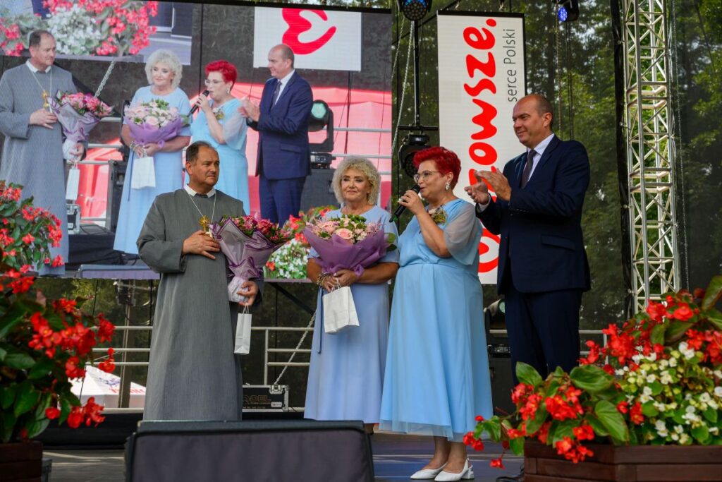 Na scenie dwie kobiety w niebieskich sukienkach i dwóch mężczyzn wręczają kwiaty. Tło to ekran z napisem. Wokół sceny czerwone kwiaty.