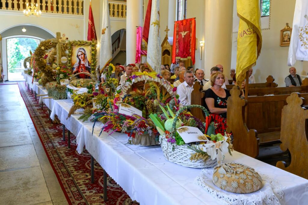 Na zdjęciu widać wnętrze kościoła z drewnianymi ławkami. Na białym obrusie leżą kolorowe wieńce i chleb ozdobny. Tło zdobią jasne ściany i kolorowe flagi.