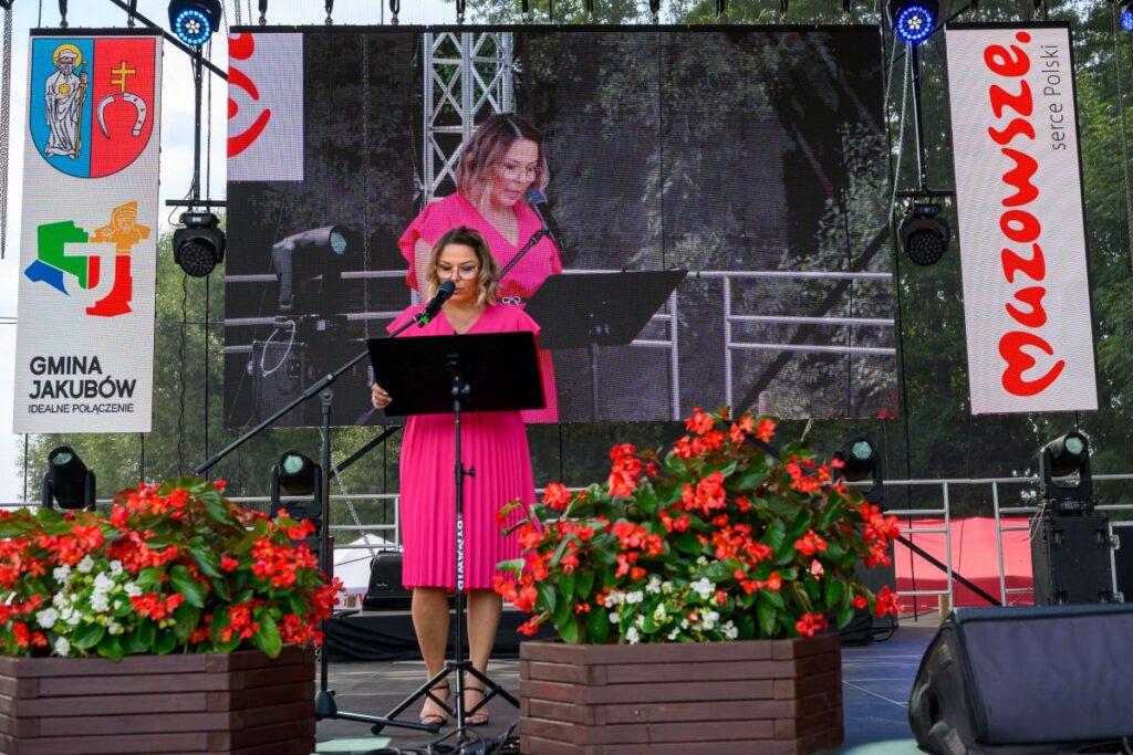 Kobieta w różowej sukience przemawia na scenie z mikrofonem. Tło stanowi duży ekran i kwiaty w donicach. Obok są bannery z nazwą Mazowsze.