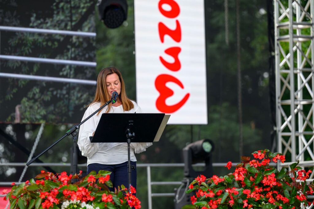 Kobieta w białej bluzce przemawia przy mikrofonie na scenie. W tle widać zielone drzewa i biały baner z czerwonym napisem. Przed nią rosną czerwone kwiaty begonia.