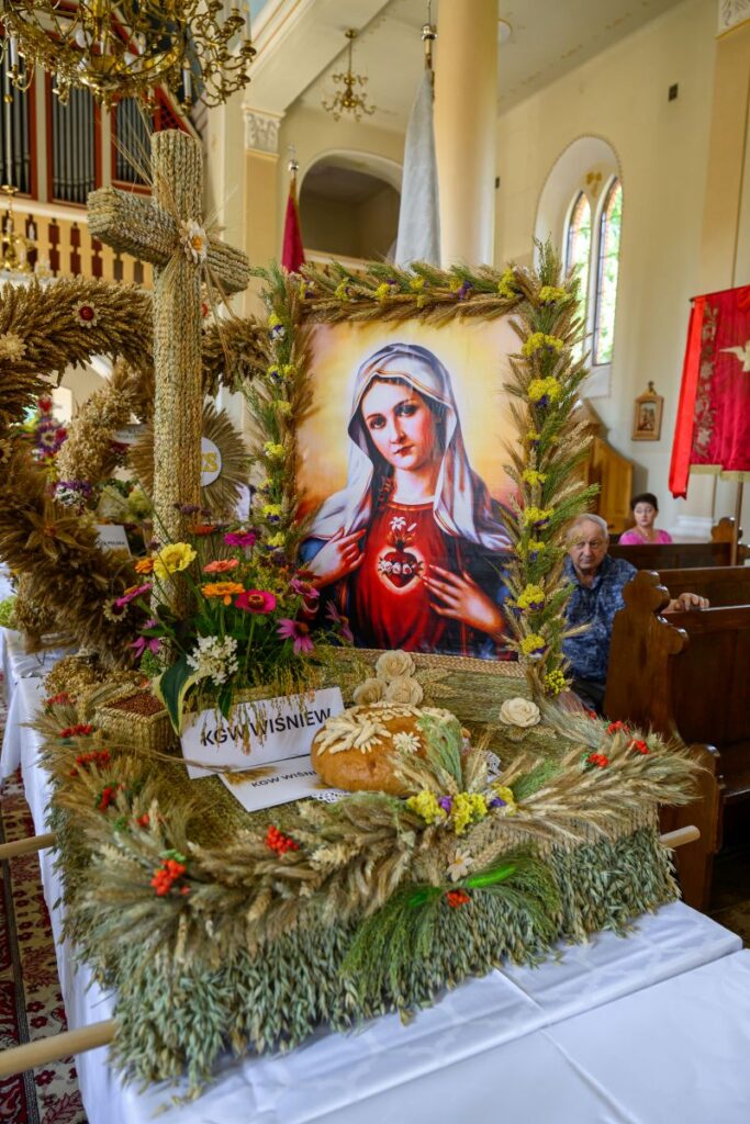 Dożynkowy wieniec z portretem Matki Boskiej otoczony zbożami i kwiatami. W tle kościół z jasnymi ścianami. Góra ozdobiona krzyżem ze słomy.
