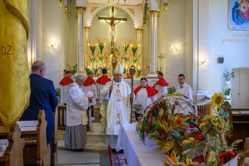 Wnętrze kościoła z ołtarzem zdobionym kwiatami. Duchowni w białych i czerwonych szatach. Przed nimi kosz z plonami.