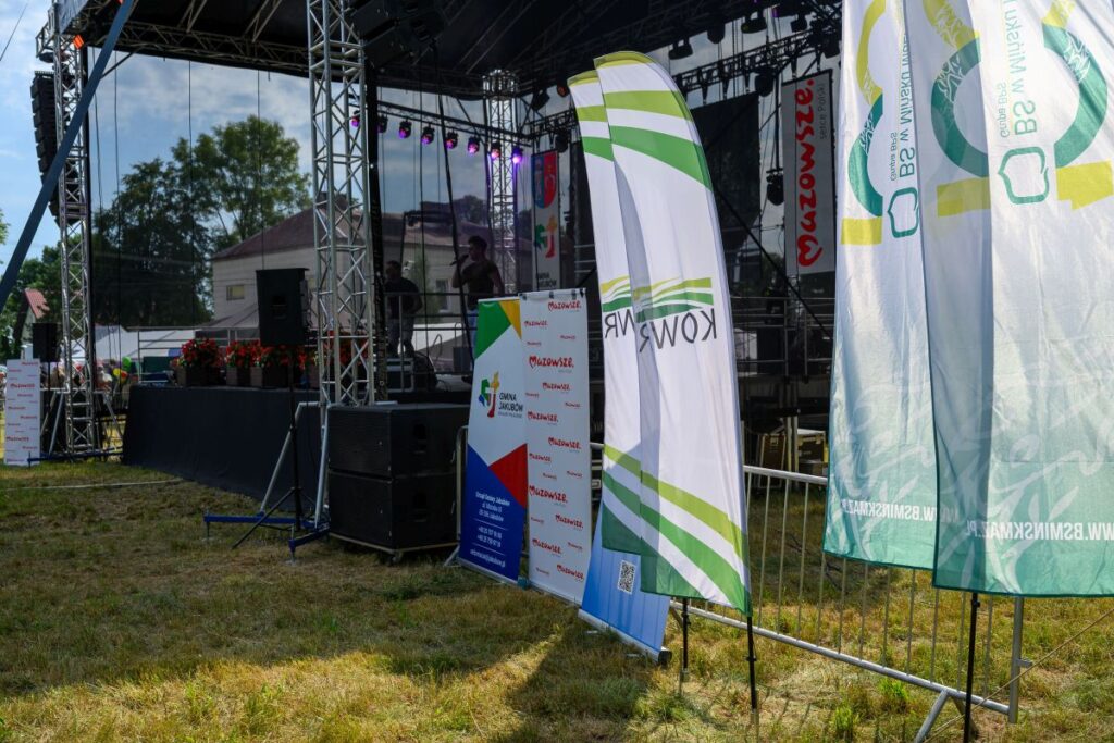 Scena plenerowa z nagłośnieniem, stoją flagi regionalne i reklamy. Tło to zieleń drzew i niebieskie niebo. Dzień słoneczny, atmosfera festiwalowa.