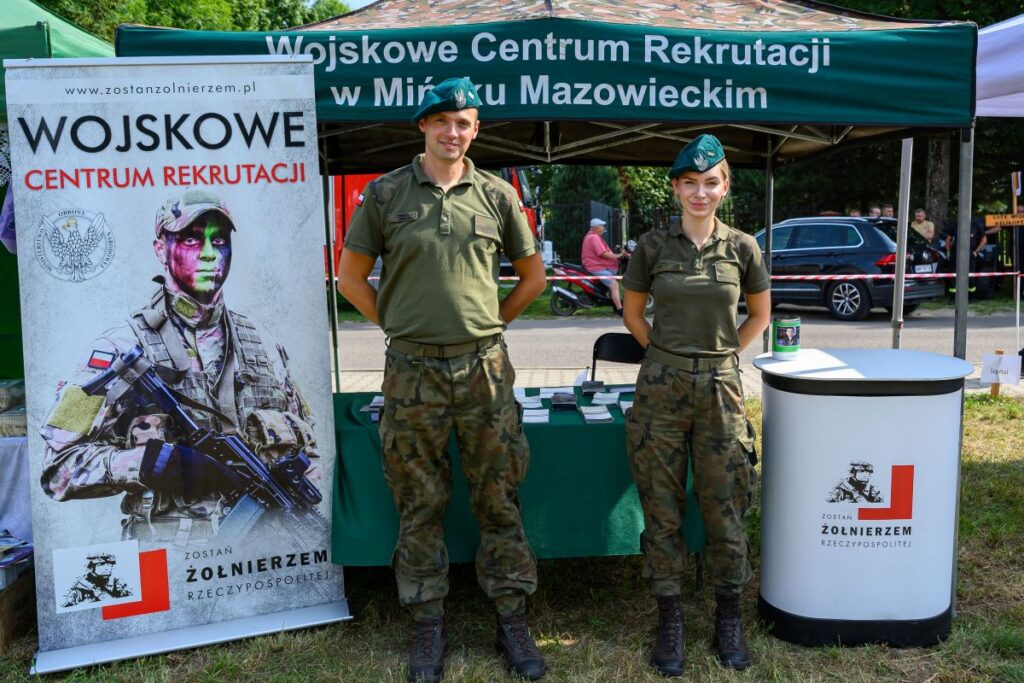 Zielony namiot rekrutacyjny z żołnierzami w mundurach stoi na trawie. Obok plakat promocyjny z wojskowym w kamuflażu. W tle park i samochód.