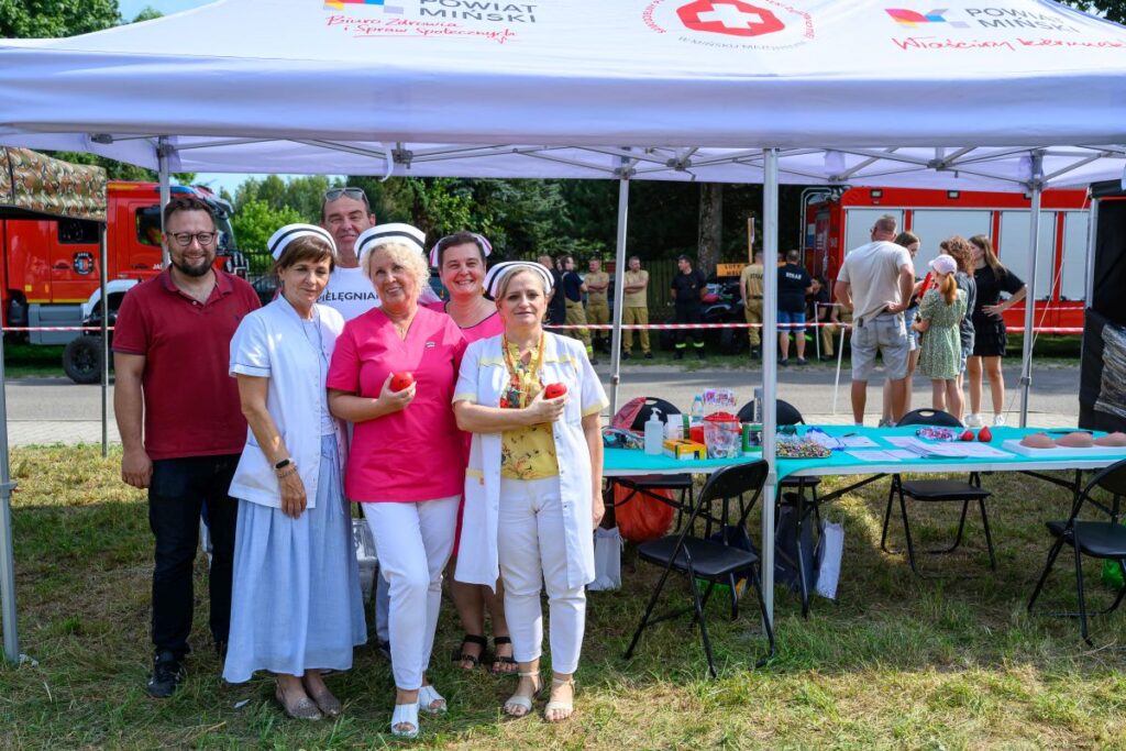 Ludzie w strojach medycznych i cywilnych pod białym namiotem. Obok stolik z materiałami, w tle wóz strażacki. Słoneczny dzień.