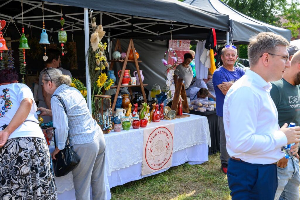 Stoisko z ceramiką i ozdobami na festynie; kolorowe przedmioty, słoneczniki; czarne namioty na tle zieleni.