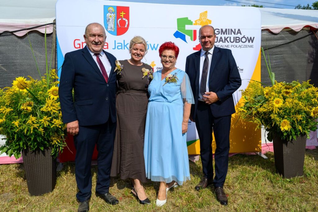 Cztery osoby w eleganckich strojach pozują na tle banera z logo gminy Jakubów. Obok stoją ozdobne donice z żółtymi kwiatami. Tło z zieloną trawą i niebieskim niebem.