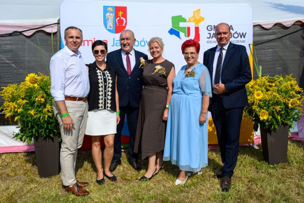 Na zdjęciu sześć osób przed białym banerem z logotypem gminy. Mają formalne ubrania, wokół są żółte kwiaty. Tło to zielone pole.