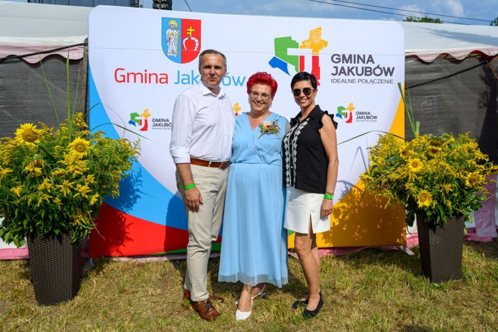 Trzy osoby stoją przed kolorowym banerem z logiem Gmina Jakubów i ozdobnymi żółtymi kwiatami na trawie.