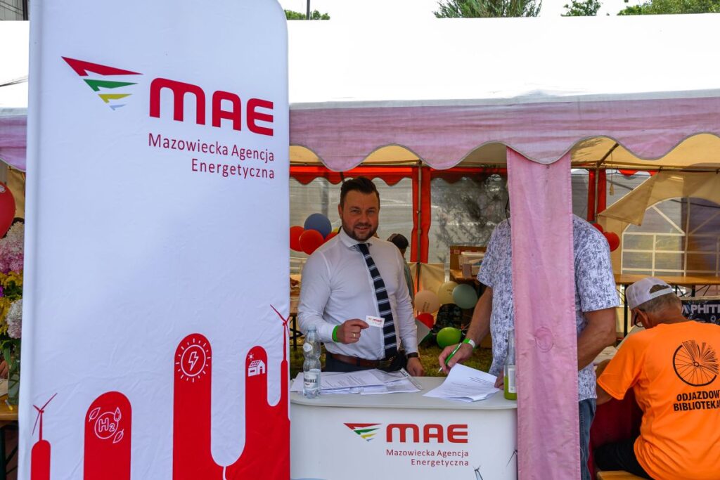 Stoisko Mazowieckiej Agencji Energetycznej z białym i czerwonym banerem. Mężczyzna w białej koszuli stoi przy stole z ulotkami. W tle kolorowe balony i namiot.