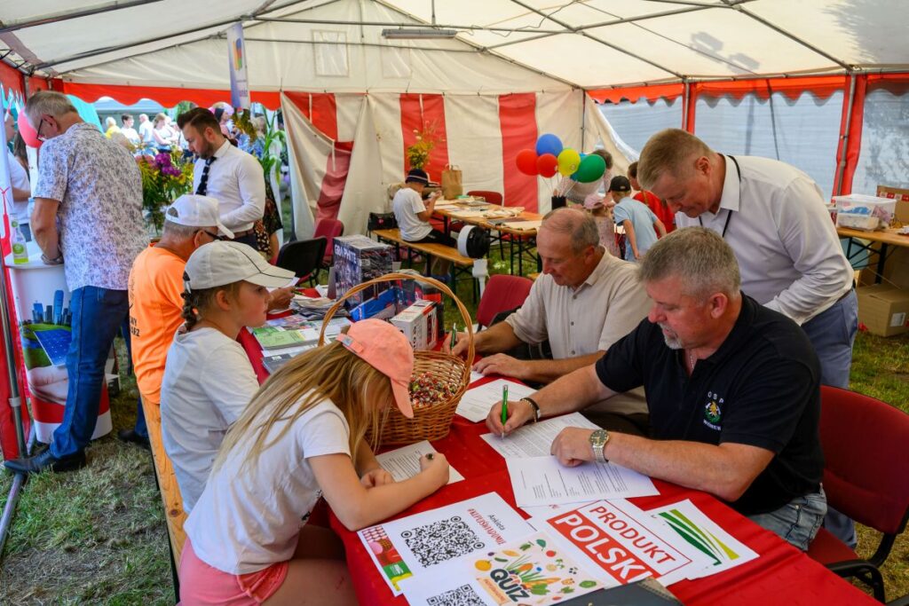 Na zdjęciu ludzie siedzą przy stołach w namiocie, na czerwonym obrusie są dokumenty i kosz z cukierkami. Tło ozdobione balonami i plakatem o produkcie polskim. Namiot jest biało-czerwony, a uczestnicy zajęci są pisaniem