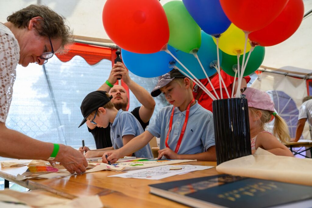 Dzieci rysują na drewnianym stole pod białym namiotem. Obok stoją czerwone, niebieskie i zielone balony. W tle osoby dorosłe pomagają dzieciom.