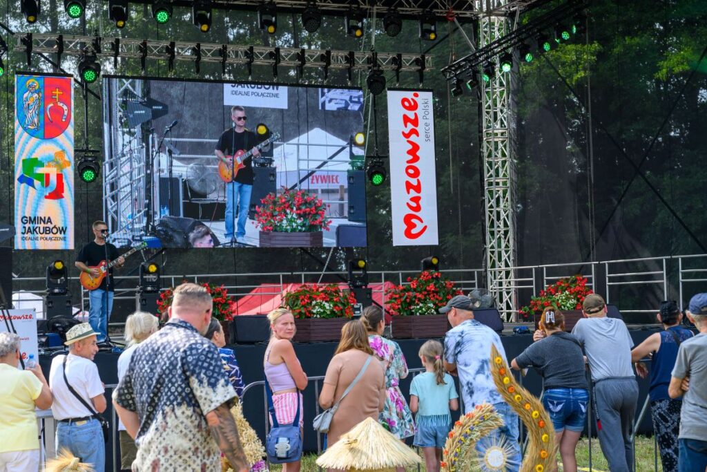 Koncert plenerowy z gitarzystą na scenie; kolorowe logo i kwiaty tła. Publiczność ogląda występ na dziedzińcu.