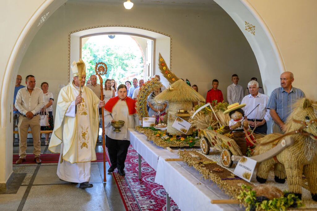 Na zdjęciu widoczna jest procesja religijna w kościele. Osoba w stroju liturgicznym prowadzi grupę ludzi, a przed nimi są ozdoby z słomy i zbóż na białym stole. Tło stanowi jasne pomieszczenie z łukowym wejściem.