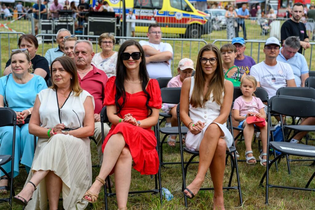 Grupa ludzi siedzi na krzesłach na zewnątrz, niebo jest jasne. W centrum kobiety w kolorowych sukienkach. W tle widać żółtą karetkę.